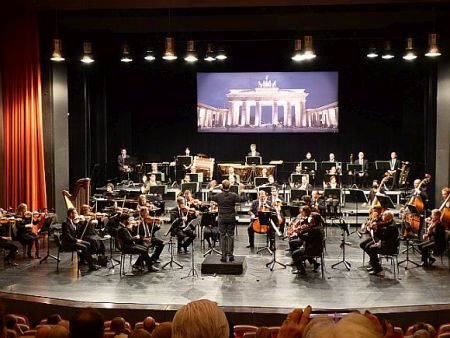 Blick auf die Bühne mit den Symphonikern und Dirigent; Foto vom Brandenburger Tor im Hintergrund